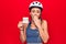 Young beautiful cyclist woman wearing bike security helmet holding reminder with help message covering mouth with hand, shocked