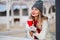 Young beautiful dreaming girl holds hot cappuccino coffee in red cup in hands with white mittens and smiles in morning