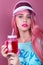 Young beautiful fashionable woman with trendy glowing neon pink hair and pink lips in sun visor drinking summer cocktail