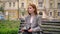 Young beautiful ginger woman in jacket sitting on bench and holding tablet, city street background