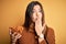 Young beautiful girl eating baked german pretzel standing over isolated yellow background cover mouth with hand shocked with shame