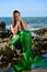 A young beautiful girl in a green mermaid costume sits on a stone on the beach against the sky, hugging her neck with her hands an