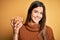 Young beautiful girl holding bowl of salty peanuts standing over yellow background with a happy face standing and smiling with a