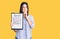 Young beautiful girl holding clipboard with agreement document serious face thinking about question with hand on chin, thoughtful