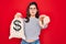 Young beautiful girl holding money bag with dollar symbol for business wealth over red background pointing with finger to the