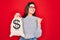 Young beautiful girl holding money bag with dollar symbol for business wealth over red background pointing and showing with thumb