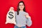 Young beautiful girl holding money bag with dollar symbol for business wealth over red background with surprise face pointing