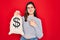 Young beautiful girl holding money bag with dollar symbol for business wealth over red background very happy pointing with hand