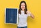 Young beautiful girl holding photovoltaic solar panel smiling happy and positive, thumb up doing excellent and approval sign