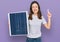 Young beautiful girl holding photovoltaic solar panel surprised with an idea or question pointing finger with happy face, number