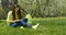 Young beautiful girl with long dark hair sitting in nature on a green field with dandelions under a tree, reading a book