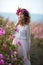 Young beautiful girl in a long dress and a wreath of flowers in the garden of lilac bush