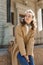 Young beautiful girl with long hair in beige coat on a sunny day sitting on a curb and smiling. Street style portrait