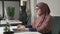 Young beautiful girl in pink hijab and glasses typing, working on computer. Look at the camera. Arab women in the office
