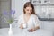 A young beautiful girl sits at a white table in the kitchen and looks at the phone, reads the morning news over a cup of coffee