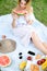 Young beautiful girl sitting on plaid near fruits and hat, eating watermelon, grass in background.