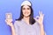 Young beautiful girl wearing sleep mask holding pills doing ok sign with fingers, smiling friendly gesturing excellent symbol