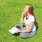 A young beautiful girl in a white T-shirt with her eyes closed sits on the green grass on the lawn with a notebook and a pen,