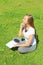 A young beautiful girl in a white T-shirt with her eyes closed sits on the green grass on the lawn with a notebook and a pen,