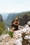 Young beautiful girl woman in black sporty slim outfit sitting on edge rock mountain cliff peak with view on valley