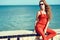 Young beautiful glam stylish woman wearing coral red jumpsuit and dark trendy sunglasses sitting on the parapet at the seaside