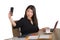 Young beautiful and happy Asian Chinese business woman taking selfie photo with mobile phone at corporate company office desk smil