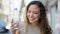 Young beautiful hispanic woman holding bottle of water smiling at street