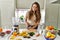 Young beautiful hispanic woman preparing vegetable smoothie with blender cutting banana at the kitchen