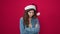 Young beautiful hispanic woman standing with unhappy expression wearing christmas hat over isolated red background