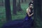 Young beautiful lady in luxurious sequin evening dress sitting in the mysterious misty woods leaning on the moss covered tree