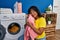 Young beautiful latin woman washing clothes touching face with soft sweater at laundry room