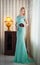 Young beautiful luxurious woman in long elegant dress. Beautiful young blonde woman in turquoise dress with curtains in background