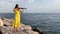 Young beautiful musician girl in yellow dress is playing electric violin on rocks near Mediterranean sea. Open air solo concert