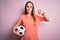 Young beautiful player woman playing soccer holding football ball  over pink background with angry face, negative sign showing