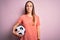 Young beautiful player woman playing soccer holding football ball  over pink background with a confident expression on smart face