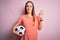 Young beautiful player woman playing soccer holding football ball  over pink background doing ok sign with fingers, excellent