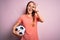 Young beautiful player woman playing soccer holding football ball  over pink background with happy face smiling doing ok sign with