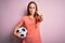 Young beautiful player woman playing soccer holding football ball  over pink background pointing with finger to the camera and to