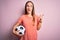 Young beautiful player woman playing soccer holding football ball  over pink background pointing and showing with thumb up to the