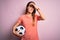 Young beautiful player woman playing soccer holding football ball  over pink background stressed with hand on head, shocked with