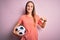 Young beautiful player woman playing soccer holding football ball  over pink background with surprise face pointing finger to