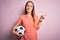 Young beautiful player woman playing soccer holding football ball  over pink background very happy pointing with hand and finger