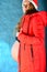 A young and beautiful, pregnant girl in a red jacket and Santa Claus hat is standing on a blue background and shows the belly