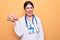 Young beautiful psychiatrist woman wearing stethoscope holding brain over yellow background looking positive and happy standing