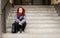 Young beautiful red hair girl sitting alone outdoors on the stairs of the building with hat and shirt feeling anxious and depresse