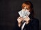 Young beautiful red-haired business woman in black formal jacket holding bundle of dollars in hands in front of her face