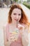 Young beautiful red-haired woman with braces drinks cooling lemonade outdoors in summer. Portrait of a smiling girl with