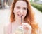 Young beautiful red-haired woman with braces drinks cooling lemonade outdoors in summer. Portrait of a smiling girl with