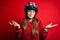 Young beautiful redhead motocyclist woman wearing moto helmet over red background clueless and confused expression with arms and