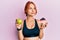 Young beautiful redhead woman holding green apple and cake slice smiling looking to the side and staring away thinking